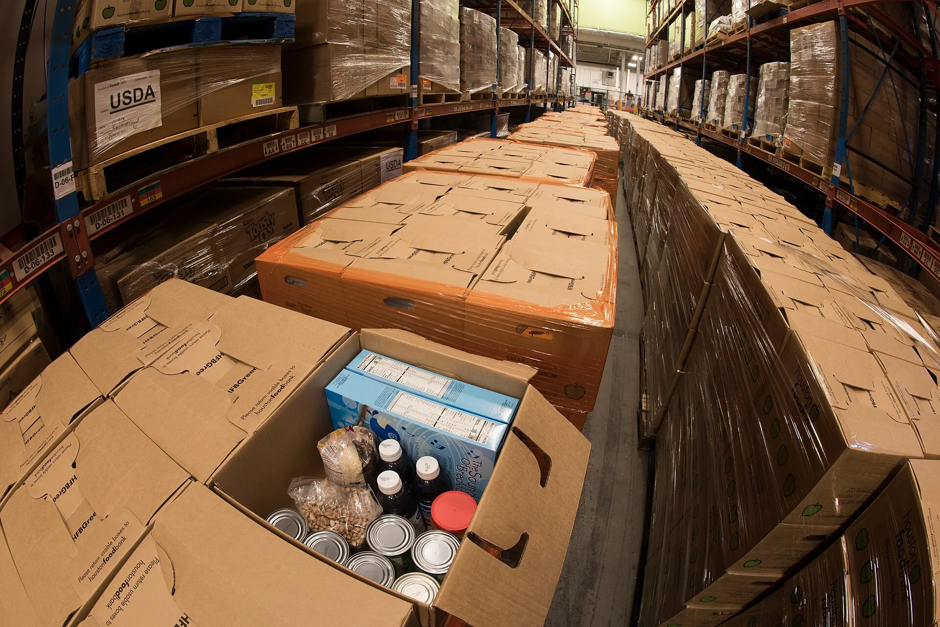 Pallets of USDA Emergency Food Assistance Program (TEFAP) commodities wait on the Houston Food Bank commodity warehouse shelves for movement to packing lines or delivery to food pantries; pallets in the aisles are loaded with disaster assistance packages (those with orange wrapping have USDA Foods i