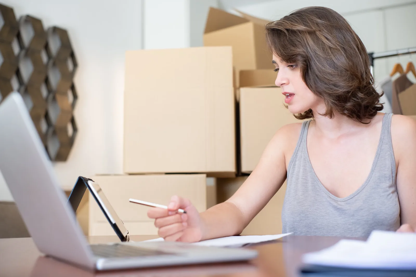 Woman running an online store, managing orders with a laptop and tablet, surrounded by cardboard boxes.