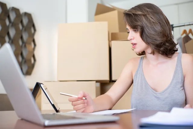Woman running an online store, managing orders with a laptop and tablet, surrounded by cardboard boxes. Marketplace seller software selection: five compliance gates
