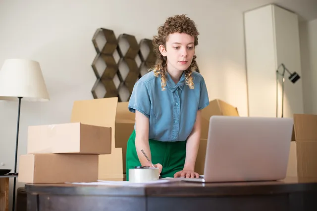 A young woman working on her laptop surrounded by cardboard boxes, indicating online business operations. Marketplace seller software UK regulations: trigger map